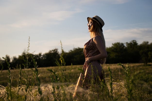 Beautiful girl in dress and hat on the field in the setting sun.