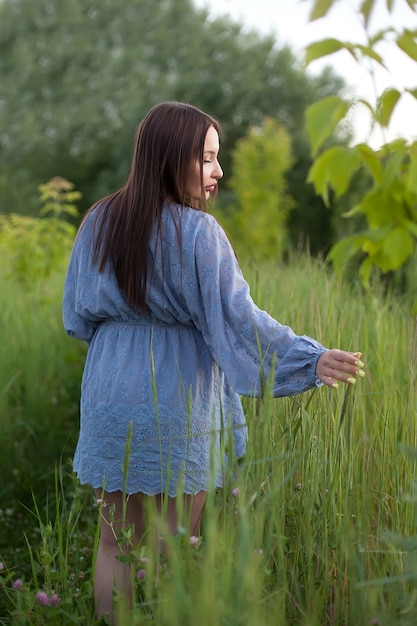 A beautiful girl in a dress walks across the field and touches the green spikelets with her hand.