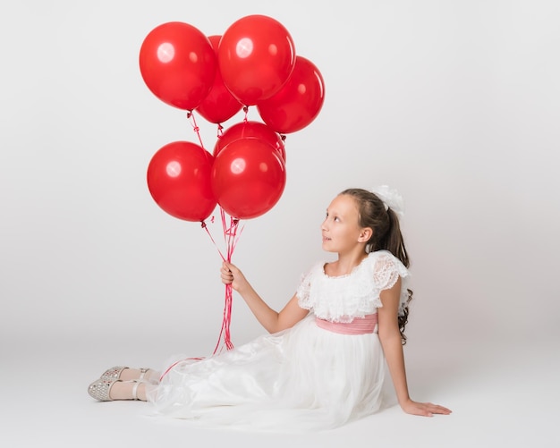 Beautiful girl dressed in long white dress holding lot of red balloons in hand looking up at balls