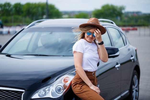 A beautiful girl of European appearance in glasses and a brown hat stands near a black car. Photoshoot near the car.