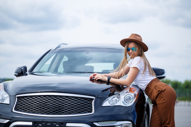 A beautiful girl of European appearance with glasses and a brown hat is standing near a black car. Photo near the car.