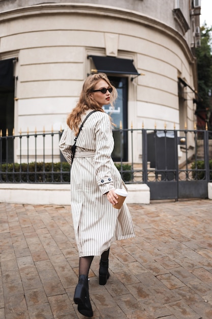 Beautiful girl from back in striped trench coat and sunglasses holding coffee to go in hand while dreamily walking around cozy city street