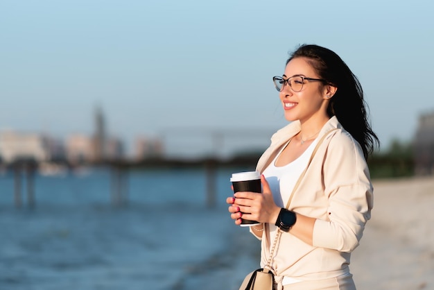 Beautiful girl in glasses stands on the beach by the river smiles holds a paper cup with a hot drink in her hands Coffee mockup