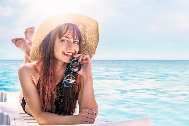 Beautiful girl in a hat and beautiful makeup lying on a lounger against beach and ocean Beautiful girl in a swimsuit with sunglasses resting on a deckchair on background of sea Fashion model