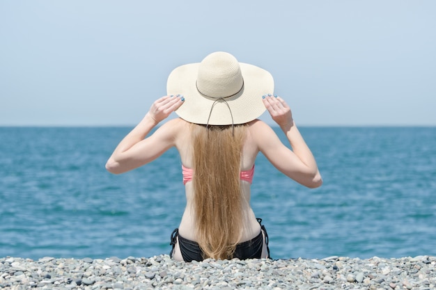 Beautiful girl in a hat and swimsuit sits on the beach. Sea on the background. View from the back