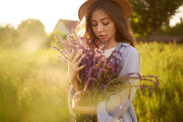 A beautiful girl in a hat with a bouquet of lavender. She is dressed in a blue shirt, a white T-shirt and a hat.