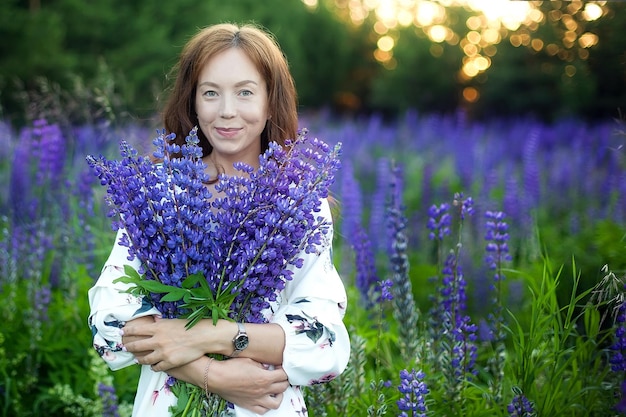 Beautiful girl in a hat with a bouquet of lupine flowers A girl in a yellow dress a straw hat with a bouquet of lupins stands in a flower field Young brunette girl Summer time Lupine field