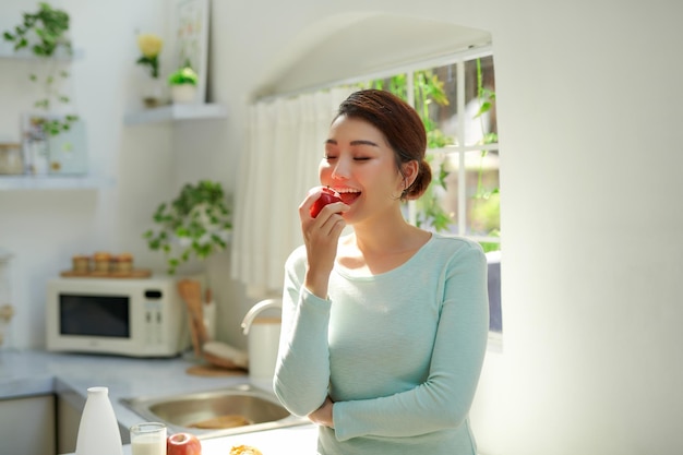 Beautiful girl holding red apple while standing in the home kitchen in the morning