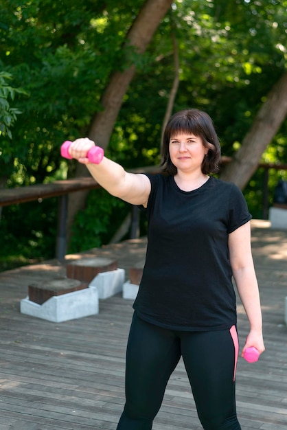 Beautiful girl is engaged in fitness in the park and looks into the distance. Pink dumbbells, shakes arm muscles