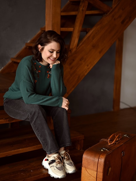 A beautiful girl is sitting next to a suitcase on a wooden staircase and looks out the window