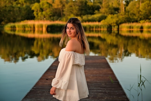 A beautiful girl is standing on the pier in the background there is a river a lake