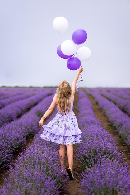 Beautiful girl in a lilac dress runs through a field with lavender In the hands of a bunch of balloons Rear view