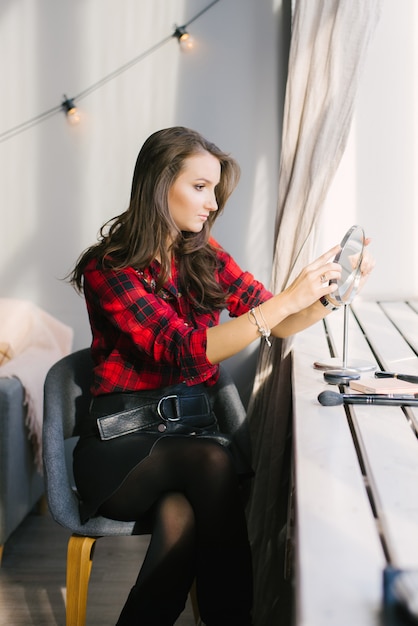 A beautiful girl looks at herself in the table mirror by the window. Going to do her makeup