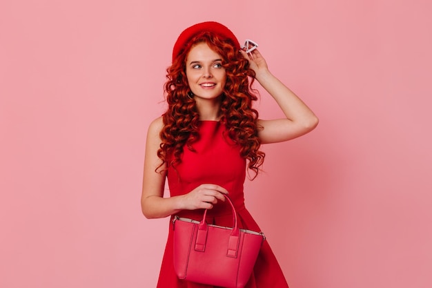 Beautiful girl in minimalistic dress posing with bright bag and touching her red beret on pink background