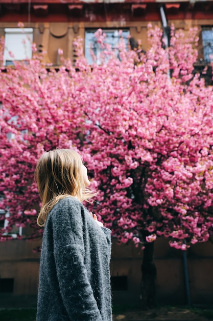 Beautiful girl near the sakura trees. Woman in dress and stylish coat Pink flowers blooming in Uzhhorod. Blossom around. Spring time. Relaxing and happiness concept.