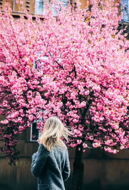 Photo beautiful girl near the sakura trees. woman in dress and stylish coat pink flowers blooming in uzhhorod. blossom around. spring time. relaxing and happiness concept.