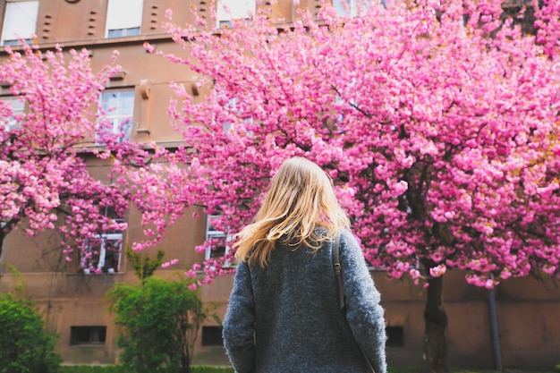 Beautiful girl near the sakura trees Woman in dress and stylish coat Pink flowers blooming in Uzhhorod Blossom around Spring time Relaxing and happiness concept