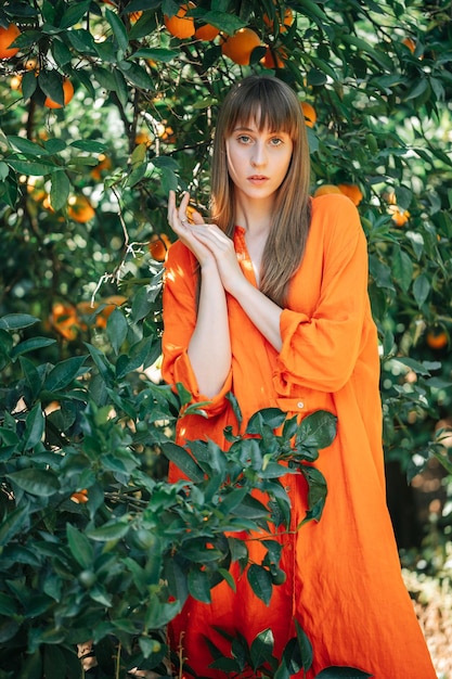 Beautiful girl in orange dress is looking at camera by holding hands together up in orange garden