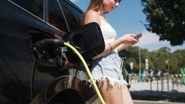 Beautiful girl plugging a cable charger into her parked black electric car and using a smartphone