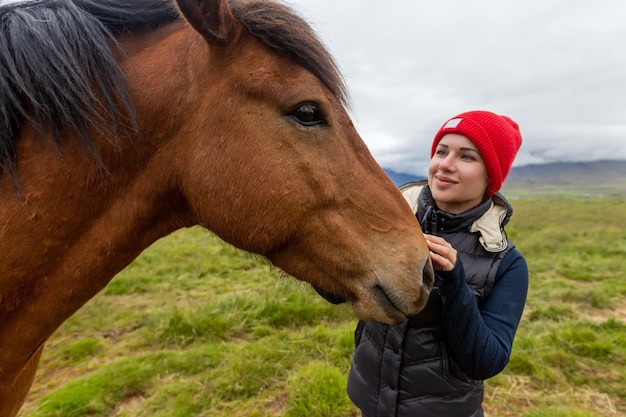 A beautiful girl in a red hat is looking at an Icelandic horse. Iceland