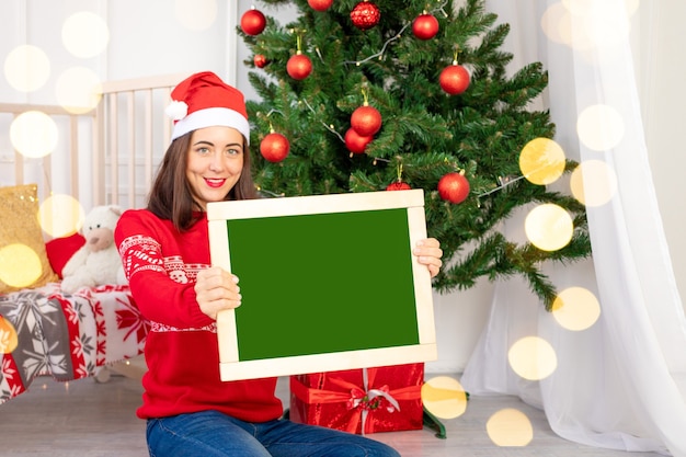 A beautiful girl in a red sweater in the children's room near the Christmas tree with a new year's interior holds a Board with space for text