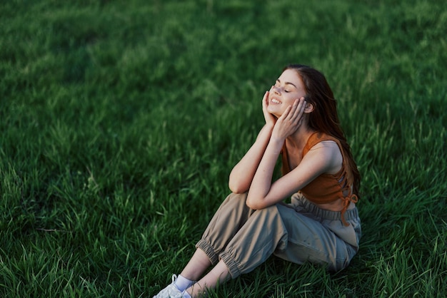 A beautiful girl relaxing in nature sitting in pants and top on the green grass in the rays of the setting summer sun The concept of health and care for the body and the environment