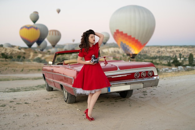 Beautiful girl retro style posing near vintage red cabriolet car with old film camera on background of balloons in Cappadocia