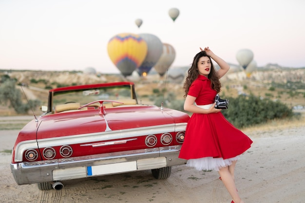 Beautiful girl retro style posing near vintage red cabriolet car with old film camera on background of balloons in Cappadocia