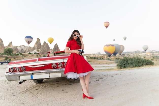 Beautiful girl retro style posing near vintage red cabriolet car with old film camera on background of balloons in Cappadocia