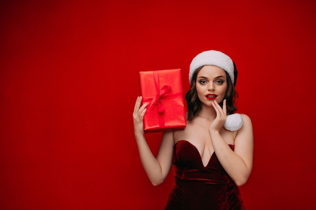 A beautiful girl in a Santa hat holds a New Years gift in her hands on a red background