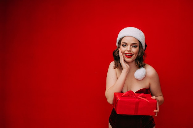 A beautiful girl in a Santa hat holds a New Years gift in her hands on a red background