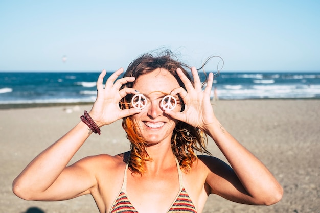 Beautiful girl showing her symbol of the peace on her eyes holded with her hands - woman in bikini at the beach smiling and looking at the camer with the sun in her face