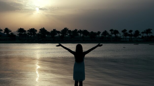 Beautiful girl silhouette standing arms wide open at sand beach in sunrise