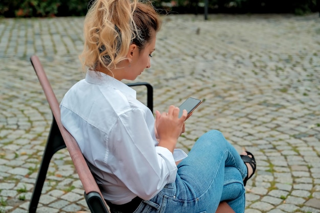 A beautiful girl sits on a bench and holds a phone in her hands the girl orders food through the phone the girl is talking on the phone laughing happy surprised