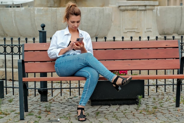 A beautiful girl sits on a bench and holds a phone in her hands the girl orders food through the phone the girl is talking on the phone laughing happy surprised