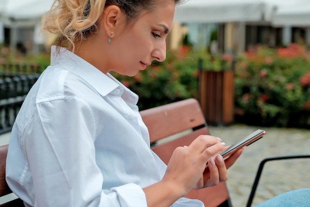 A beautiful girl sits on a bench and holds a phone in her hands the girl orders food through the phone the girl is talking on the phone laughing happy surprised