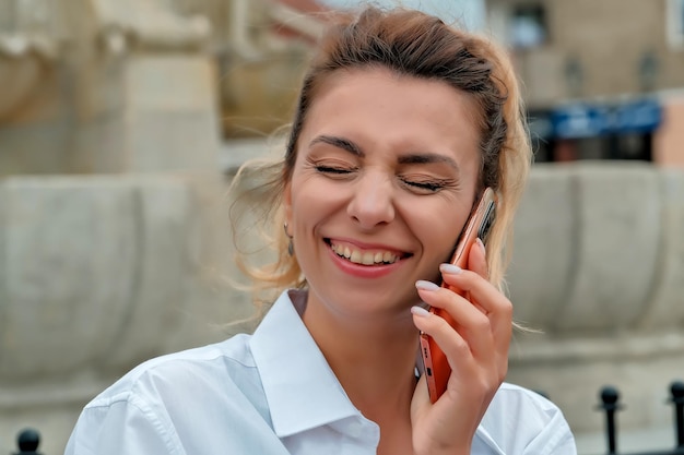 A beautiful girl sits on a bench and holds a phone in her hands the girl orders food through the phone the girl is talking on the phone laughing happy surprised