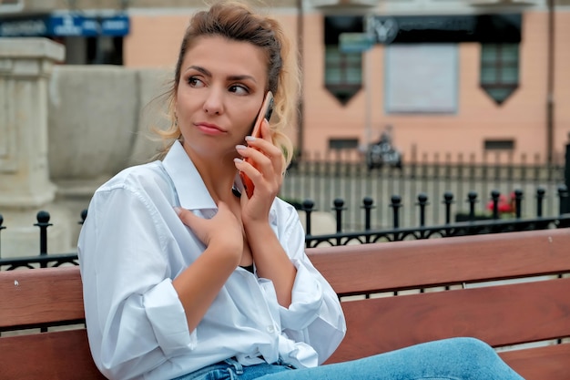 A beautiful girl sits on a bench and holds a phone in her hands the girl orders food through the phone the girl is talking on the phone laughing happy surprised