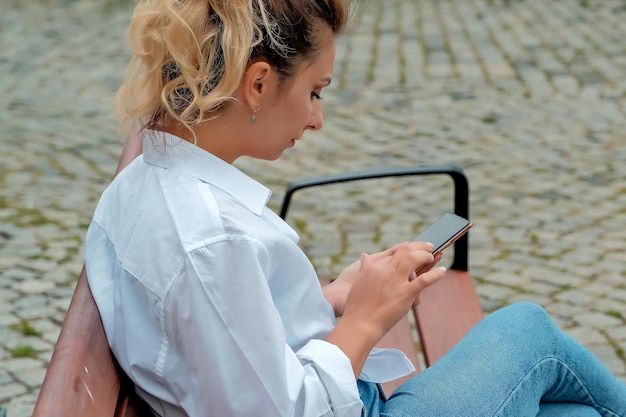 A beautiful girl sits on a bench and holds a phone in her hands the girl orders food through the phone the girl is talking on the phone laughing happy surprised