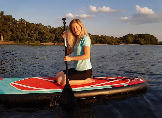 Beautiful girl sits on a SUP paddleboard and rowing an oar on the background of the river and forest