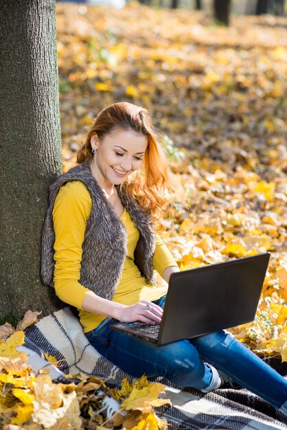 Beautiful girl sitting under a tree among the yellow leaves and uses a laptop in the autumn park