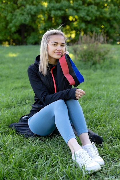 A beautiful girl in sportswear with fitness rubber bands in her hands sits on the grass