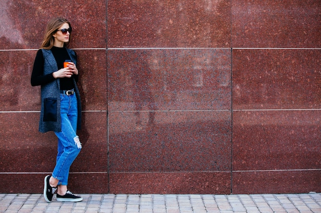 Beautiful girl stands against the wall with a coffee on the street.