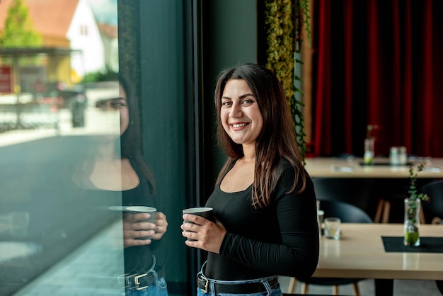 Beautiful girl stands near the window with coffee and smiles