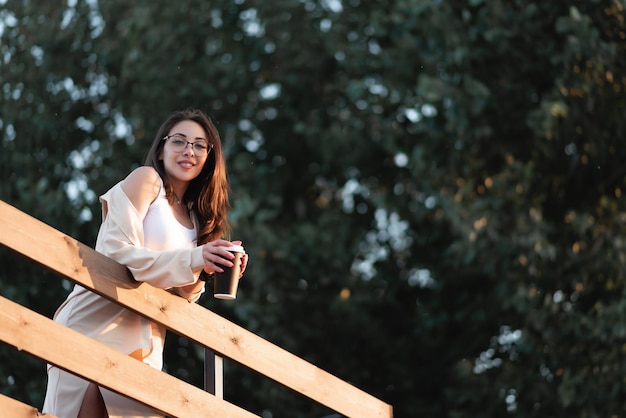 Beautiful girl stands on the veranda of a summer cafe at sunset He holds a paper cup with a hot drink in his hands Smiling looking at the camera coffee mockup