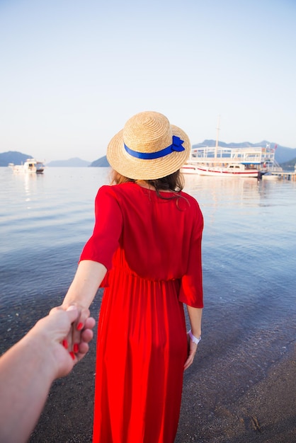 A beautiful girl in a straw hat and red dress is standing on the sea and holding her loved one's hand Romance happiness love