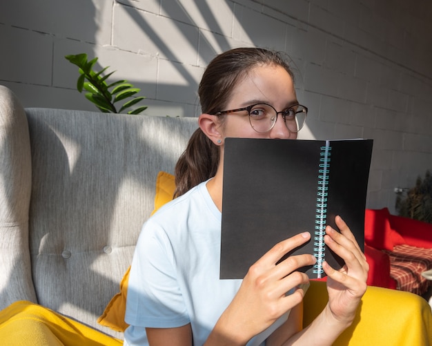 Beautiful girl student reading a book while sitting on an armchair in a cafe with stylish diagonal shadows, hard light. Concept of reading paper books. Back to school concept. Freelance concept