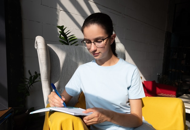 Beautiful girl student writes with a pen an assignment in a notebook while sitting on an armchair in a cafe with stylish diagonal shadows, hard light. Back to school concept. Freelance concept