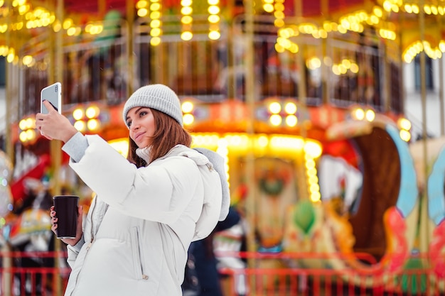 Beautiful girl takes selfie or photographs during christmas holidays while holds coffe to go in hand