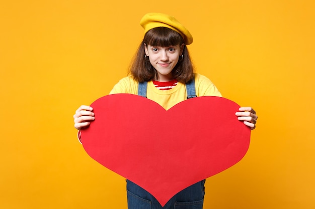 Beautiful girl teenager in french beret, denim sundress holding big empty blank red heart isolated on yellow wall background in studio. People sincere emotions, lifestyle concept. Mock up copy space.